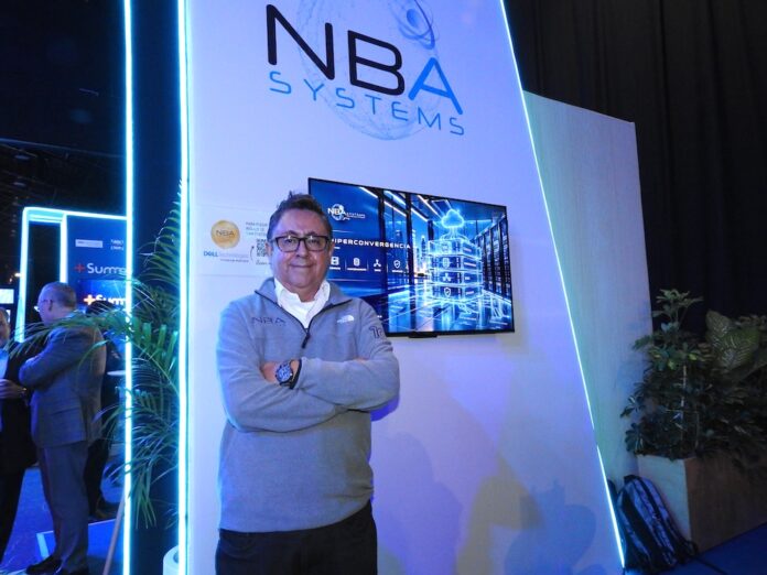 Marco González destaca la especialización de NBA Systems en almacenamiento masivo e inteligencia artificial Fotografía en plano medio de Marco González, Director de NBA Systems, vistiendo un suéter gris corporativo bordado sobre una camisa blanca, posando con los brazos cruzados frente al estand con pantalla interactiva y logotipo iluminado de la empresa.