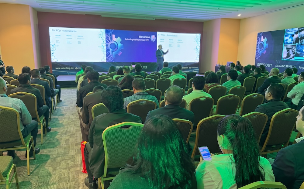 Fotografía en plano general del auditorio con los asistentes de espalda observando la presentación de Marco Teno de HPE en el escenario iluminado con tonos verdes.