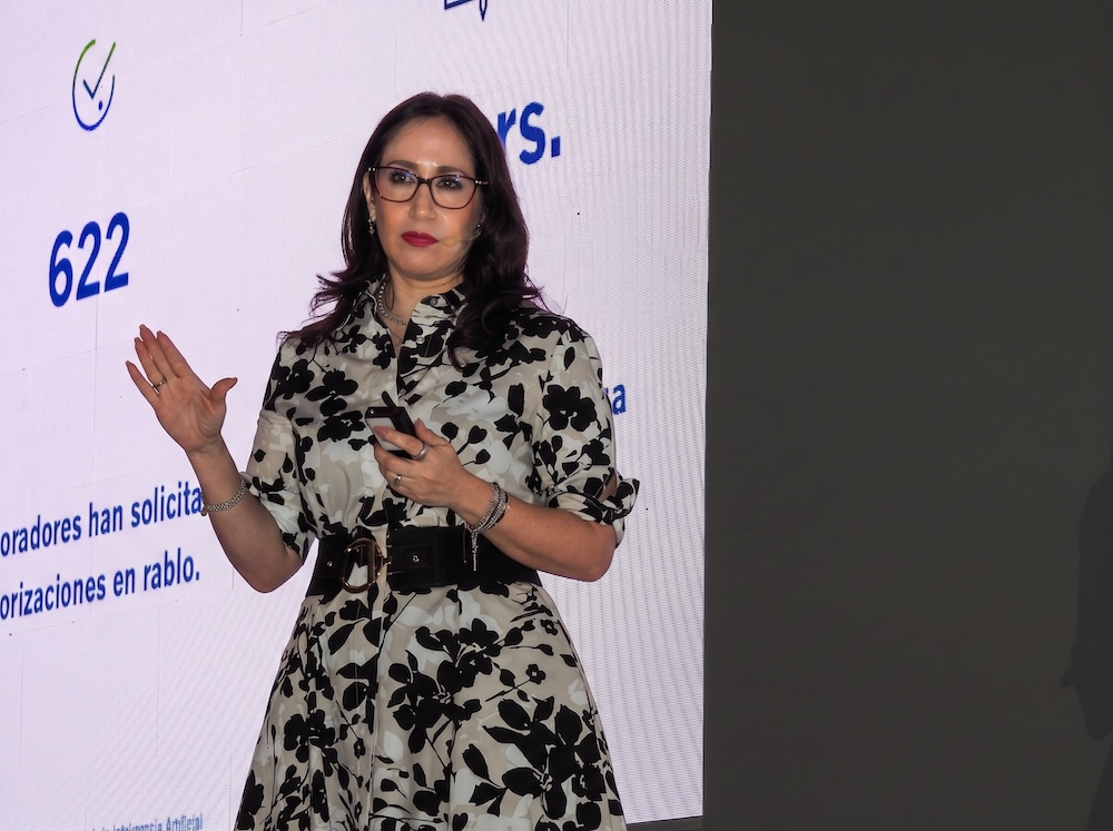 Fotografía en plano medio de Maru Moreno, Directora Comercial en CompuSoluciones, vistiendo un vestido estampado blanco y negro con gafas, exponiendo en el escenario con un micrófono de solapa.
