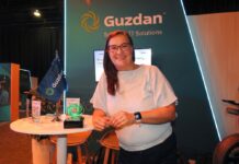 Guzdan transforma su rol como canal de distribución mediante alianzas y consultoría estratégica Fotografía en plano medio de Nadia Guzmán, Directora de Alianzas y Proyectos en Guzdan, vistiendo blusa blanca y anteojos, apoyada sonriente en una mesa alta de atención junto al trofeo conmemorativo y la bandera corporativa de Guzdan.
