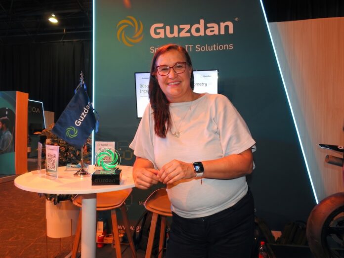 Fotografía en plano medio de Nadia Guzmán, Directora de Alianzas y Proyectos en Guzdan, vistiendo blusa blanca y anteojos, apoyada sonriente en una mesa alta de atención junto al trofeo conmemorativo y la bandera corporativa de Guzdan.