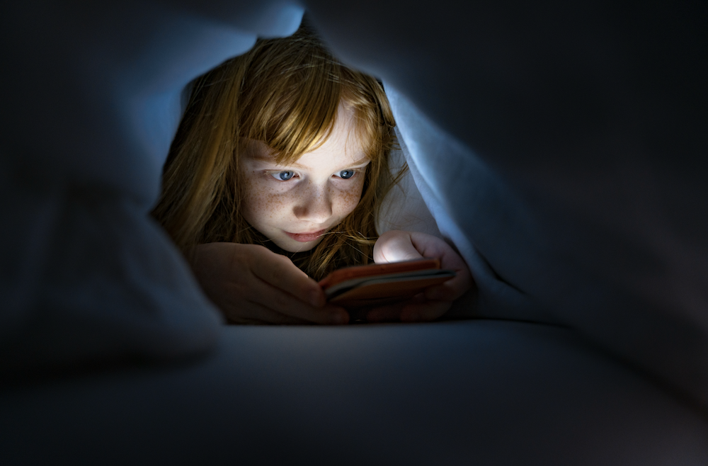 Fotografía en primer plano de una niña pelirroja recostada bajo las cobijas en la oscuridad, iluminada en el rostro únicamente por la luz reflejada de la pantalla de su teléfono inteligente.