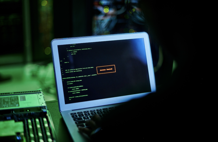 Fotografía en ángulo dorsal de un usuario frente a la pantalla de una computadora portátil en una habitación oscura con iluminación verde, donde se observan líneas de código de programación y una ventana de alerta roja central con el texto 