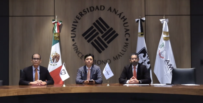 Fotografía en plano general de la mesa presidium con José Manuel Santiago, Ernesto Ibarra y Eugenio Valle sentados frente al gran escudo grabado en madera de la Universidad Anáhuac México, flanqueados por la bandera nacional y estandartes institucionales.