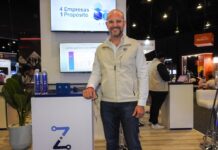 Everlay Group y la fórmula tecnológica para escalar negocios sin fronteras Fotografía en plano medio de Rodrigo Monroy, Chief Marketing Officer de Everlay Group, sonriendo con anteojos y chaleco corporativo beige de pie junto al mostrador de la plataforma Zorya, frente a una pantalla interactiva con la frase "4 Empresas 1 Propósito".