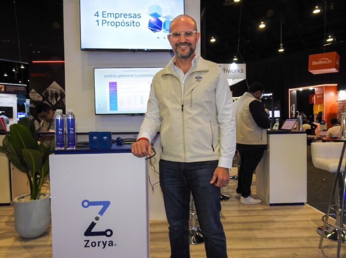 Rodrigo Monroy presenta la oferta integral de Everlay Group para optimizar procesos comerciales y fiscales Fotografía en plano medio de Rodrigo Monroy, Chief Marketing Officer de Everlay Group, sonriendo con anteojos y chaleco corporativo beige de pie junto al mostrador de la plataforma Zorya, frente a una pantalla interactiva con la frase