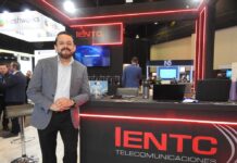 IENTC redefine la conectividad empresarial con agilidad y expansión global Fotografía en plano medio de Rubén Vargas, Manager del segmento corporativo de IENTC, vistiendo un saco a cuadros gris sobre camisa blanca y pantalón negro, posando de pie con las manos entrecruzadas frente al mostrador de exhibición de IENTC Telecomunicaciones con bordes de luz neón roja.