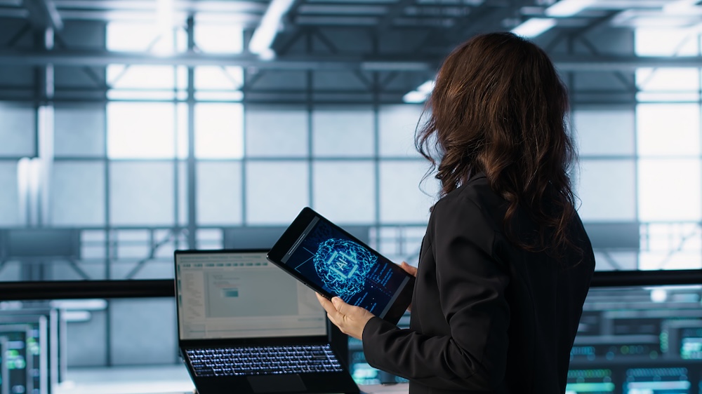 Fotografía de espaldas de una profesional corporativa de pie con traje oscuro dentro de un centro de datos o instalaciones de cómputo, sosteniendo una tableta digital donde se muestra la animación brillante de un procesador con el logotipo central "AI".
