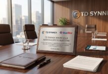 TD SYNNEX acuerda la adquisición de BlueStar para impulsar sus soluciones tecnológicas Fotografía de una placa conmemorativa metálica sobre una mesa de madera en una sala de juntas corporativa con vista panorámica a la ciudad y el logotipo de TD SYNNEX al fondo, donde se lee el anuncio de adquisición de BlueStar fechado en octubre de 2026.